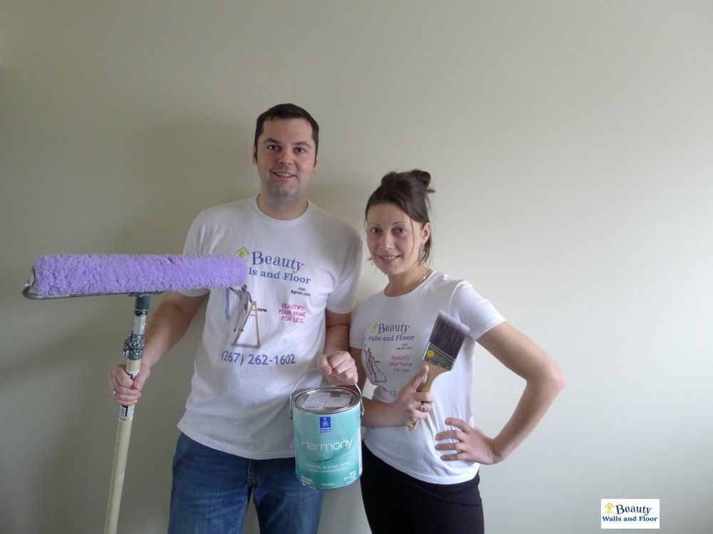 Two individuals posing with painting supplies, one holding a paint roller and the other a paint can, both wearing promotional t-shirts.