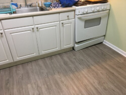 An image showing a kitchen corner with white cabinetry, a stainless steel sink, and a white stove, set against a gray wood-patterned flooring.