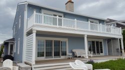 A two-story blue house with a white balcony and deck, featuring outdoor furniture and a cloudy sky background.